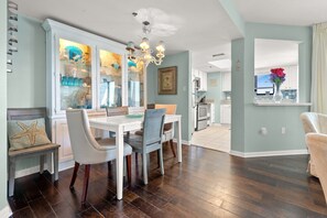 A dining area with a white table and chairs, a chandelier, and a view of the kitchen and living room through an open door. - Superb oceanfront condo w/pool, sundeck, private balcony near pier for fishing, walking, crabbing (North Myrtle Beach)