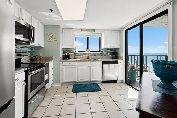 A modern kitchen with white cabinets, a built-in oven, and a dishwasher. There's a window above the sink and a view of the ocean. - Superb oceanfront condo w/pool, sundeck, private balcony near pier for fishing, walking, crabbing (North Myrtle Beach)