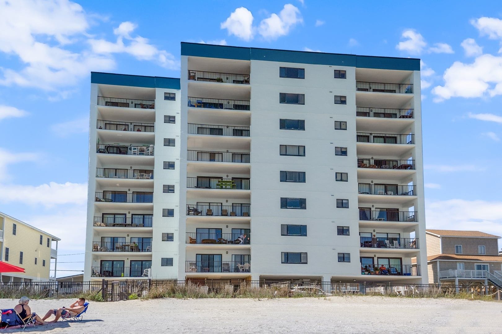 Een meerhoog hotel met balkons, een zandstrand op de voorgrond en een heldere lucht.
