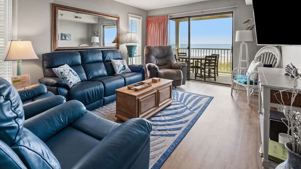 A spacious living room with a blue leather sofa, a wooden coffee table, and a dining area with a table and chairs. A large window offers a view of the ocean. - Fantastic, budget-friendly option for your next beach vacation (North Myrtle Beach)