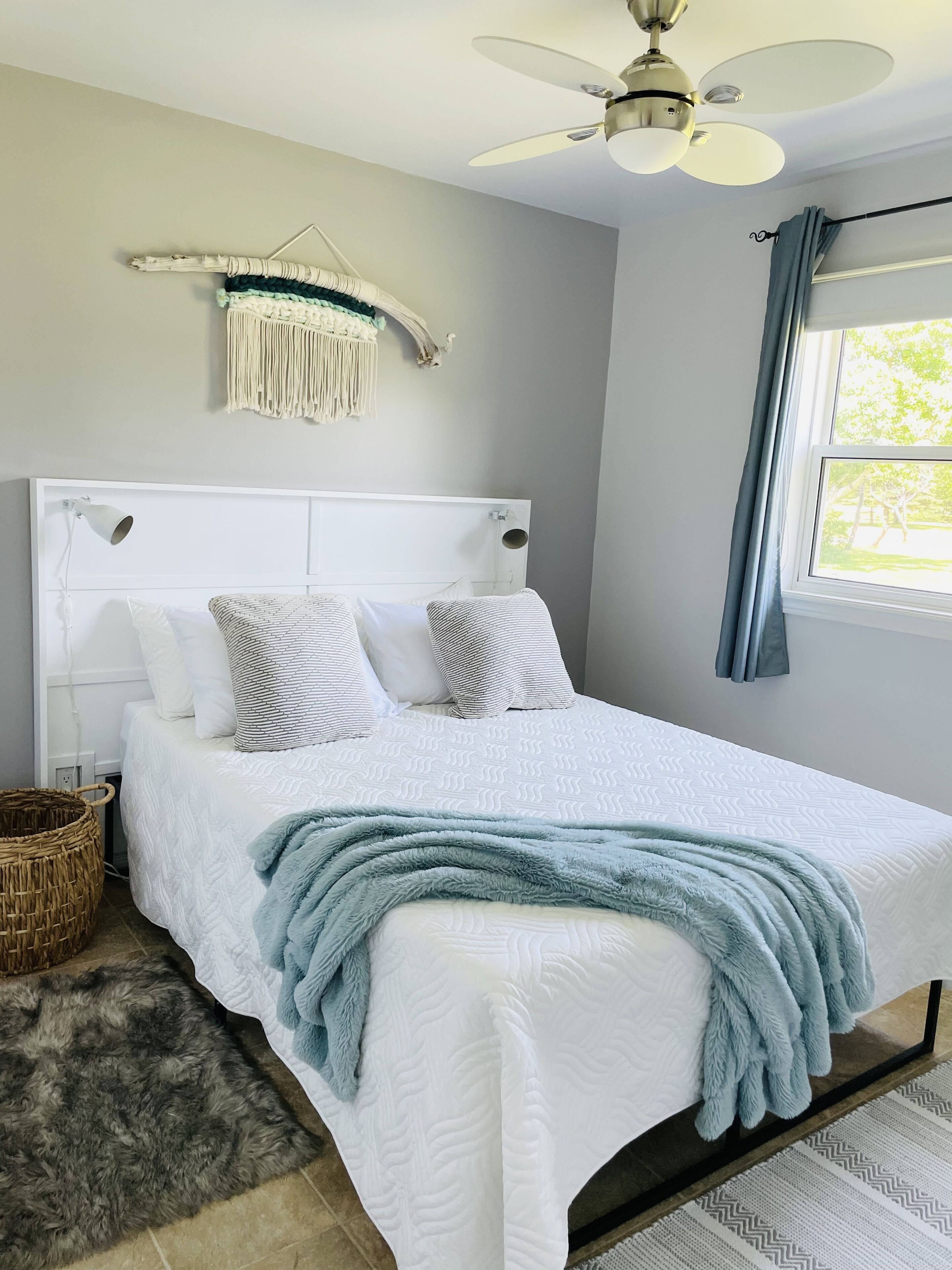 A neatly arranged bedroom with a bed, a ceiling fan, a macrame wall hanging, and a window with curtains.