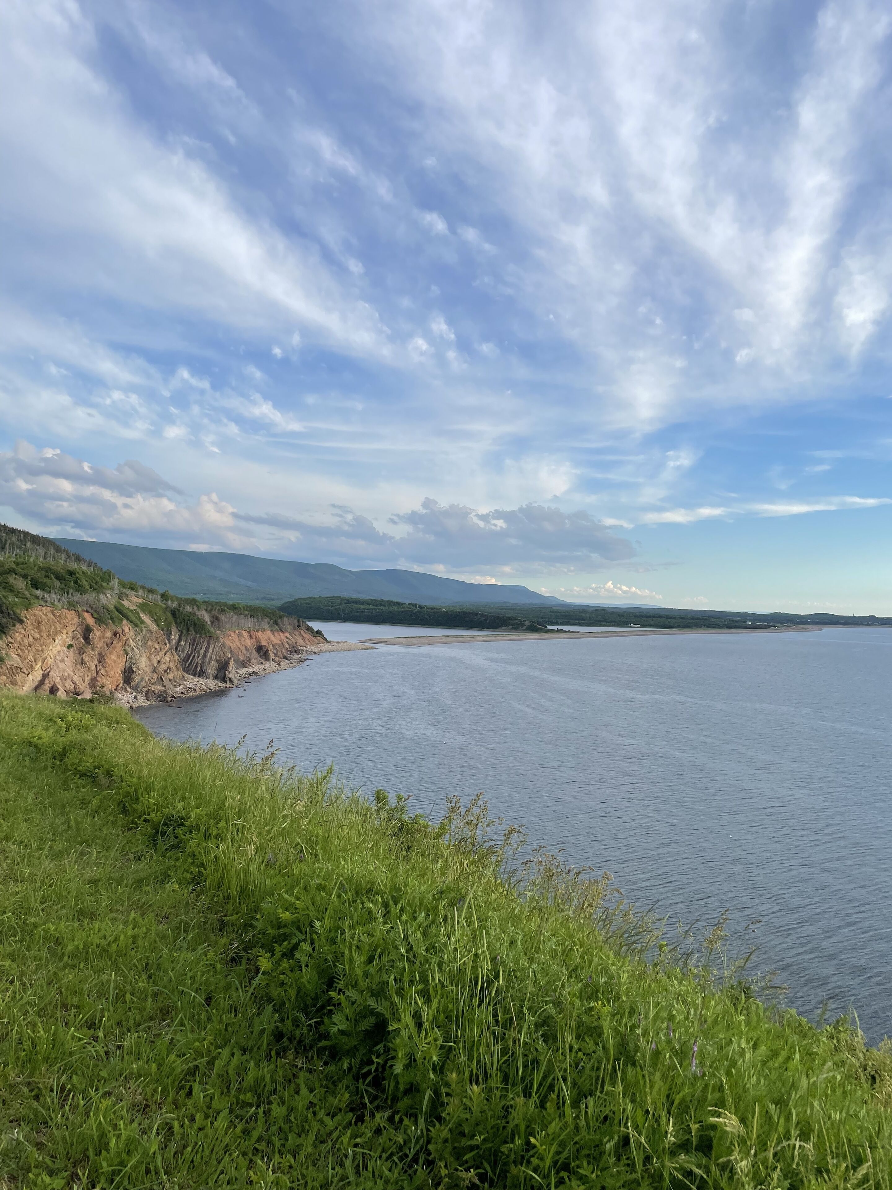 A coastal landscape with a calm body of water, grassy terrain, and distant mountains under a partly cloudy sky.