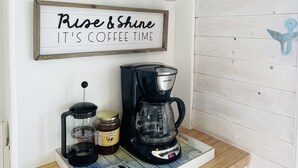Un plan de travail de cuisine avec une machine à café, un pot de sucre et un moulin à café. Au-dessus du plan de travail, on peut voir des tasses suspendues à des crochets et une pancarte sur laquelle est écrit : « Levez-vous et réveillez-vous, c’est l’heure du café ! ».