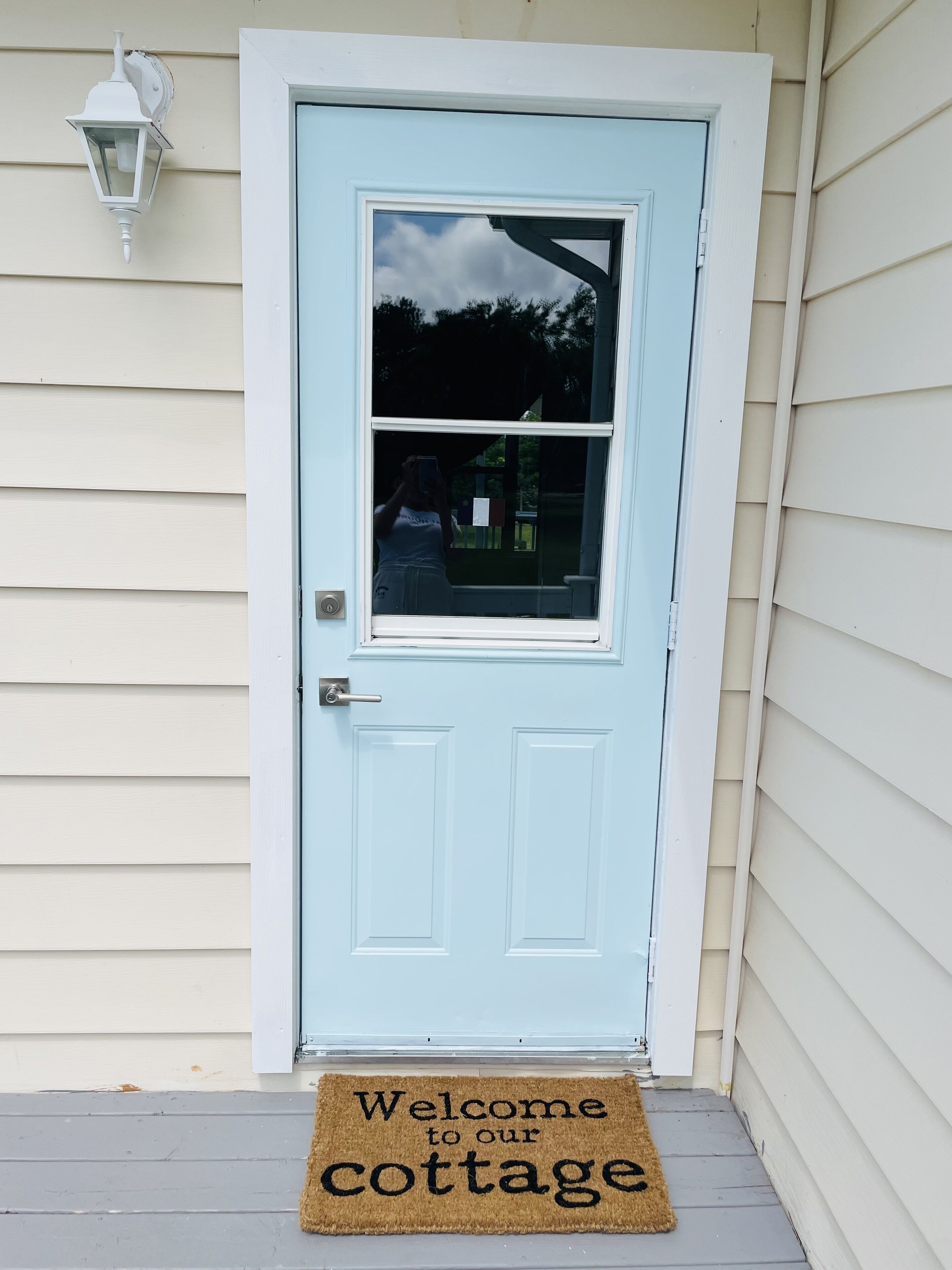A light blue door with a window, a doormat with "Welcome to our cottage" text, and a white wall-mounted light fixture.