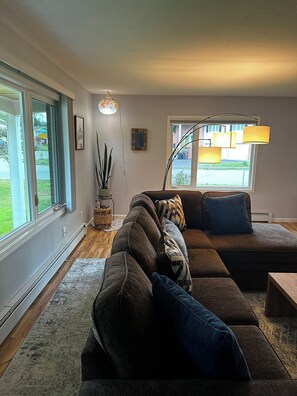 Ein modernes Wohnzimmer mit einem Ecksofa, einer Stehlampe und einem großen Fenster mit Blick ins Freie.