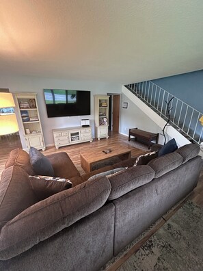 A living room with a large grey sofa, a coffee table, a flat-screen TV, and a staircase with a railing. - 3 Bedroom-Queen Beds-UMed-Stunning Mountain Views-Trails to lakes (Anchorage)