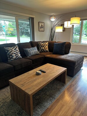 A modern living room with a sectional sofa, a wooden coffee table, and a large window providing a view of the outdoors.