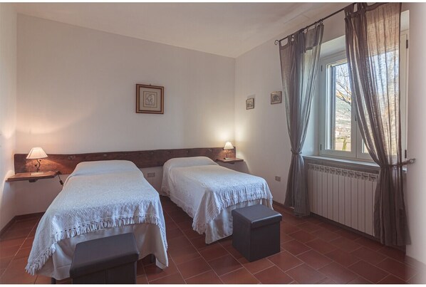 A room with two beds, a window with curtains, a radiator, and a wall-mounted picture. - OAK FOREST FARMHOUSE - LAUREL APARTMENT (Gubbio)