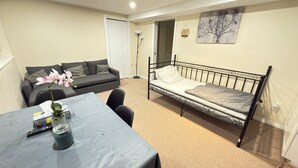 A small, well-lit room with a bed, a sofa, a table with a vase of flowers, and a painting on the wall.
