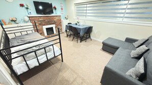 A furnished room with a bed, a sofa, a dining table with chairs, a television, and a fireplace.