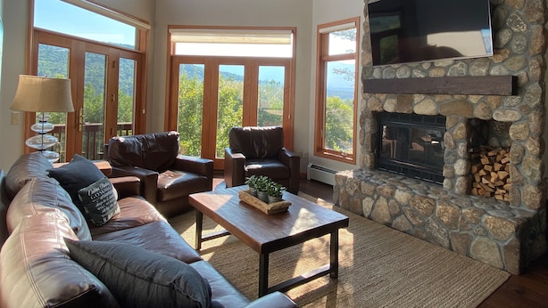 A spacious living room with leather sofas, a stone fireplace, and large windows offering a view of the outdoors. - C12 Homey Bretton Woods slopeside townhome. Pool/gym passes! (Bretton Woods)
