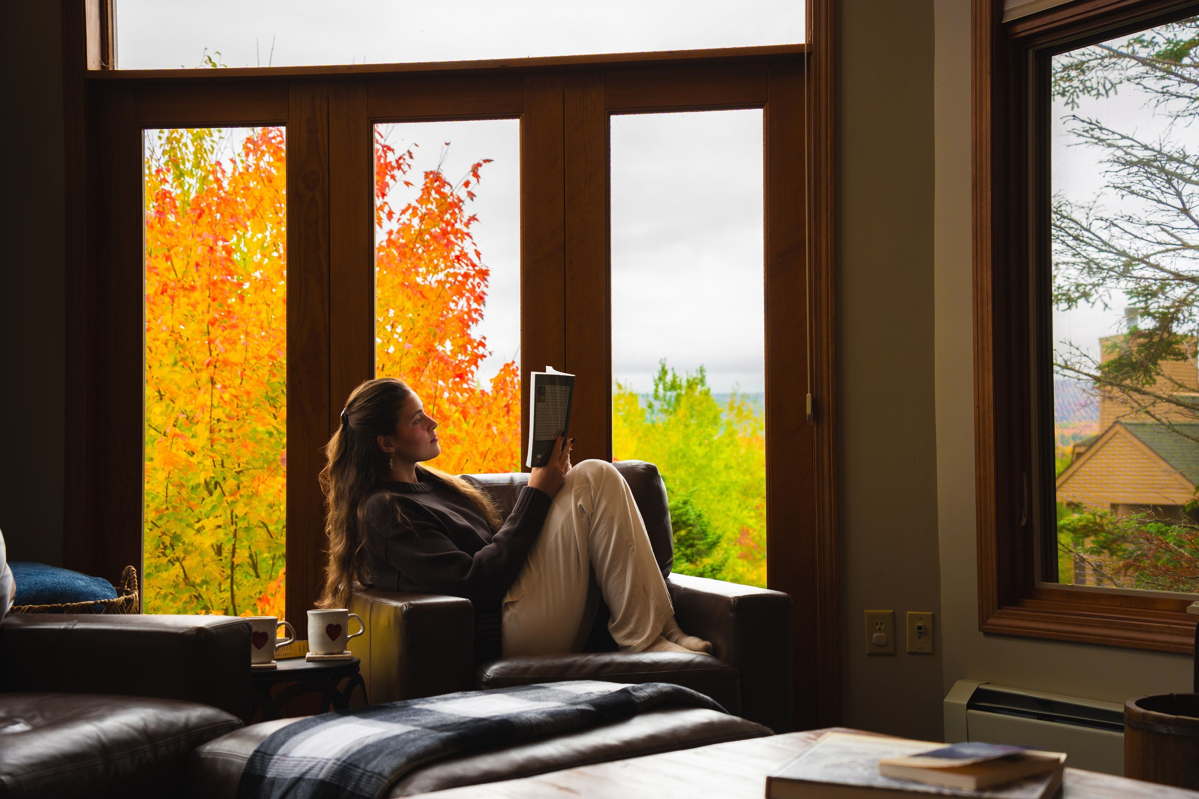 A person sitting on a couch by a window with autumn leaves outside.