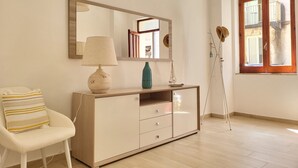 Une pièce comprenant un meuble en bois, une chaise blanche, une lampe, un miroir et une fenêtre avec des volets en bois.