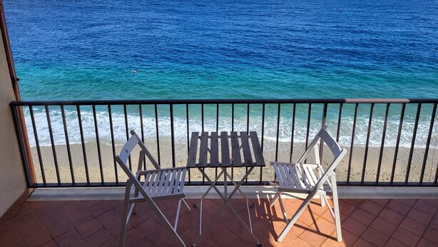 Un balcon avec deux chaises pliantes blanches et une petite table, donnant sur une plage aux eaux d’un bleu limpide et sur une côte lointaine.