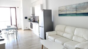 Un salon moderne avec un canapé blanc et une petite table blanche, une cuisine avec des meubles et des appareils électroménagers blancs, et un coin repas avec une table et des chaises.