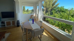 Una habitació amb taula de menjador, cadires, televisió i grans finestres amb vista a vegetació.
