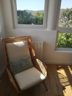 Une chaise en osier avec un coussin et un oreiller à motifs, dans une pièce dotée d’un plancher en bois, de grandes fenêtres et offrant une vue sur la verdure.