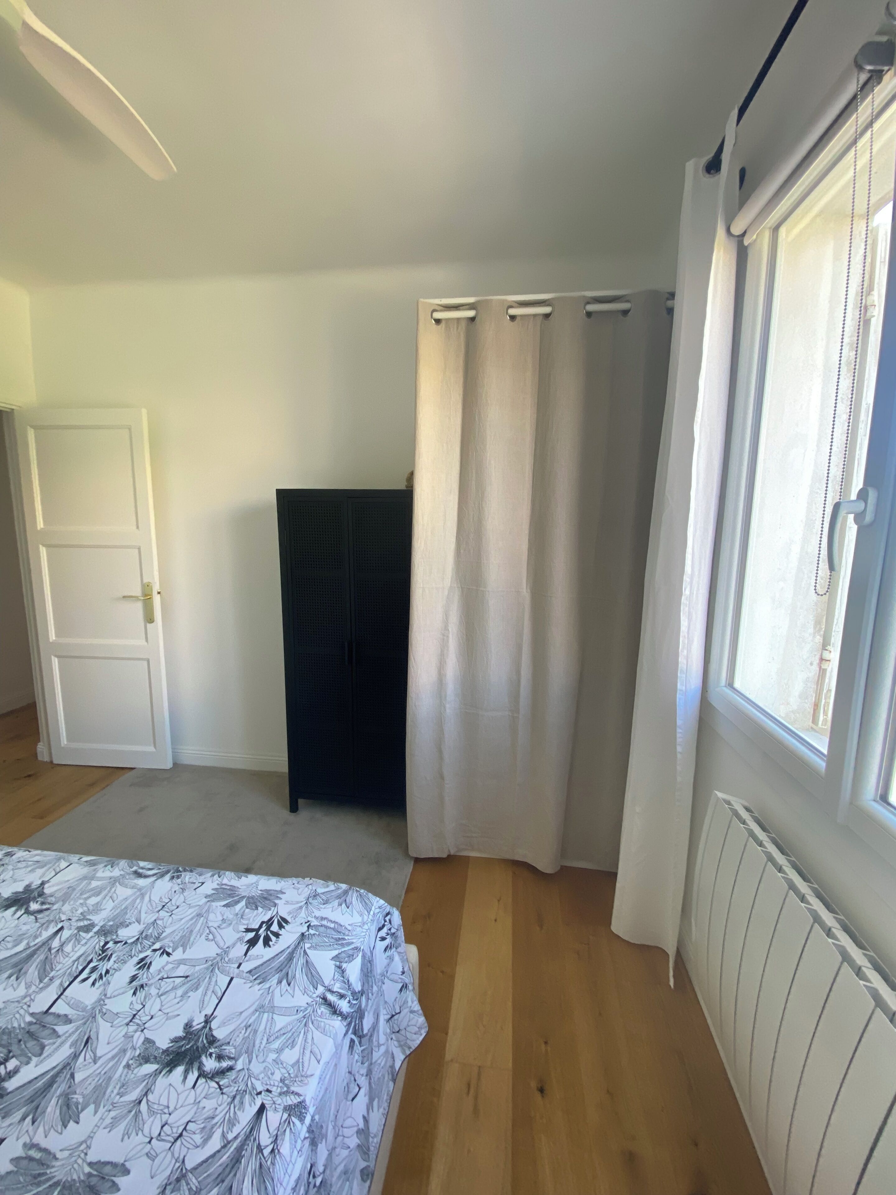 Een slaapkamer met een bed, een kledingkast, een radiator, een raam met gordijnen en een deur.