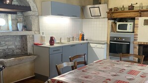 A rustic kitchen with stone walls, wooden beams, a built-in oven, and a dining area with a table covered in a patterned tablecloth. - Country house near Cabreret (Orniac)