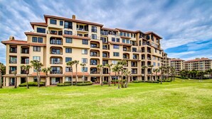 A multi-story hotel with balconies, a well-maintained lawn, and palm trees.