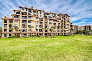 A multi-story hotel with balconies, a well-maintained lawn, and palm trees.