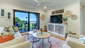 A spacious living room with a large sliding glass door leading to a balcony, a ceiling fan, and a flat-screen TV on a white cabinet.