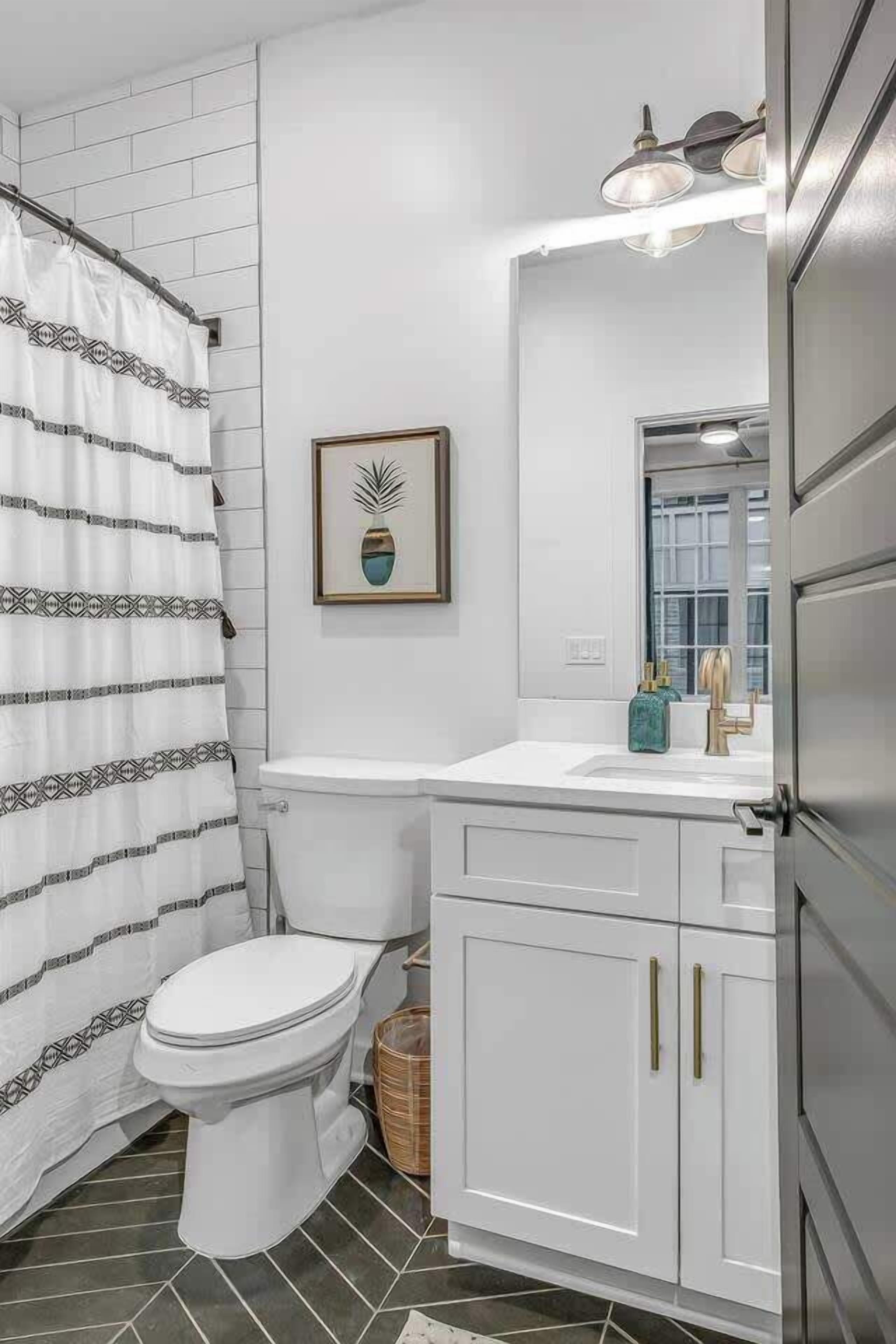 Une salle de bain avec des toilettes blanches, un rideau de douche, une plante décorative dans un cadre, un lavabo avec un miroir et un meuble.