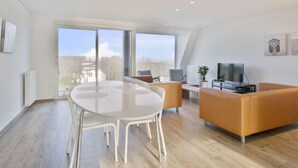 A modern living room with a round table and chairs, a sofa, a TV, and large windows offering a view of the outdoors. - L'Espérance sur mer by Interhome (Bredene)