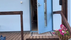 Eine Holztreppe führt zu einer blauen Tür, einer Katzenstatue und einem Blumenkorb.