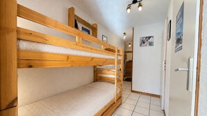 A bunk bed with a wooden frame and white bedding, a tiled floor, a door with a poster, and a hallway with a light fixture.