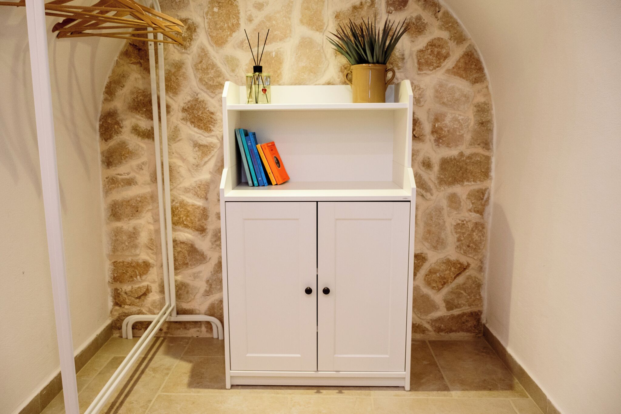 A white cabinet with two doors, a shelf with books, a potted plant, and a wall with a stone pattern.