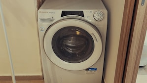 A compact, modern washing machine with a digital display, situated in a corner with a stone wall backdrop. - Nonna Vittoria Apartments, Renovated apartments in Puglia (Mola di Bari)