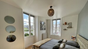 A bedroom with a bed, two windows, a radiator, and a hanging light fixture. - Le Cosy - Towncenter (Bayeux)