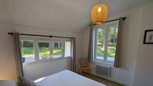 Une chambre avec un grand lit, deux fenêtres, une chaise et un luminaire suspendu.