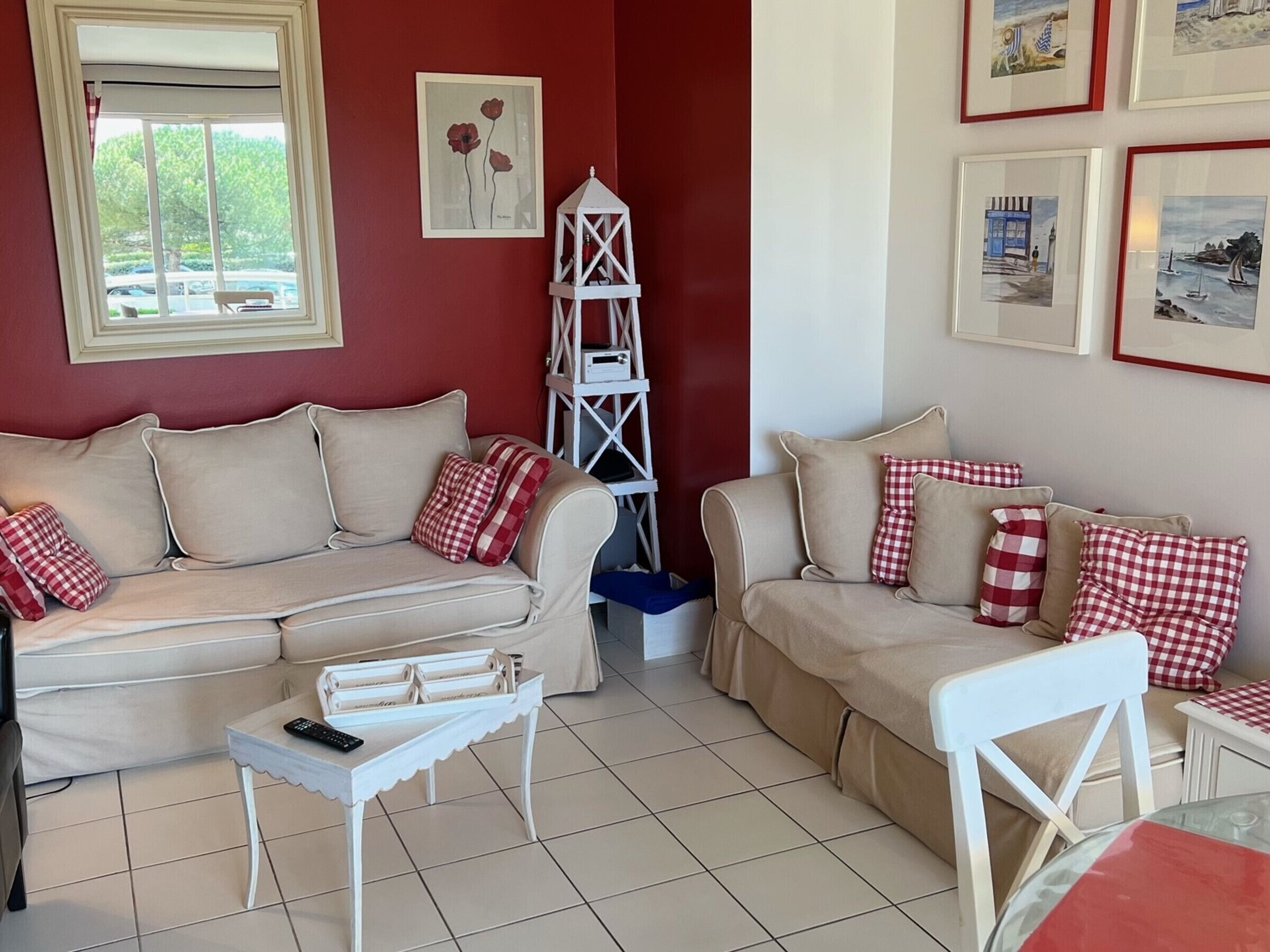 A living room with two beige sofas, red and white checkered cushions, a white side table with a remote control, and a white ladder-like shelf.