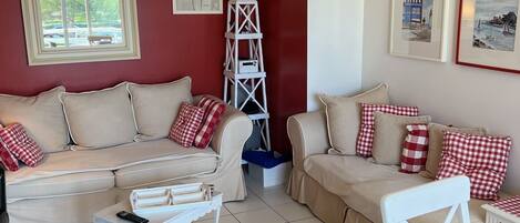A living room with two beige sofas, red and white checkered cushions, a white side table with a remote control, and a white ladder-like shelf.