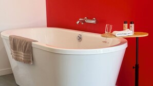 A modern bathroom with a freestanding bathtub, a wooden side table with toiletries, and a red accent wall. -  Aux Charmes (Isques)