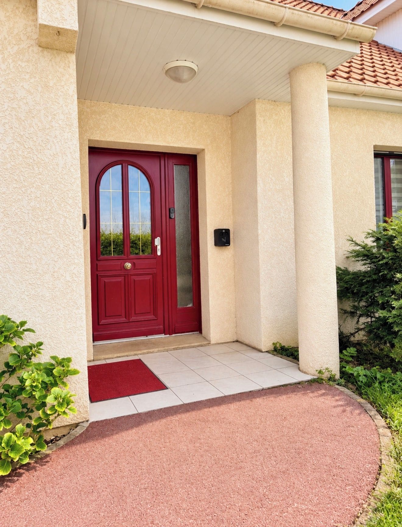 A red door with a glass panel, a white porch with a red mat, and a column.