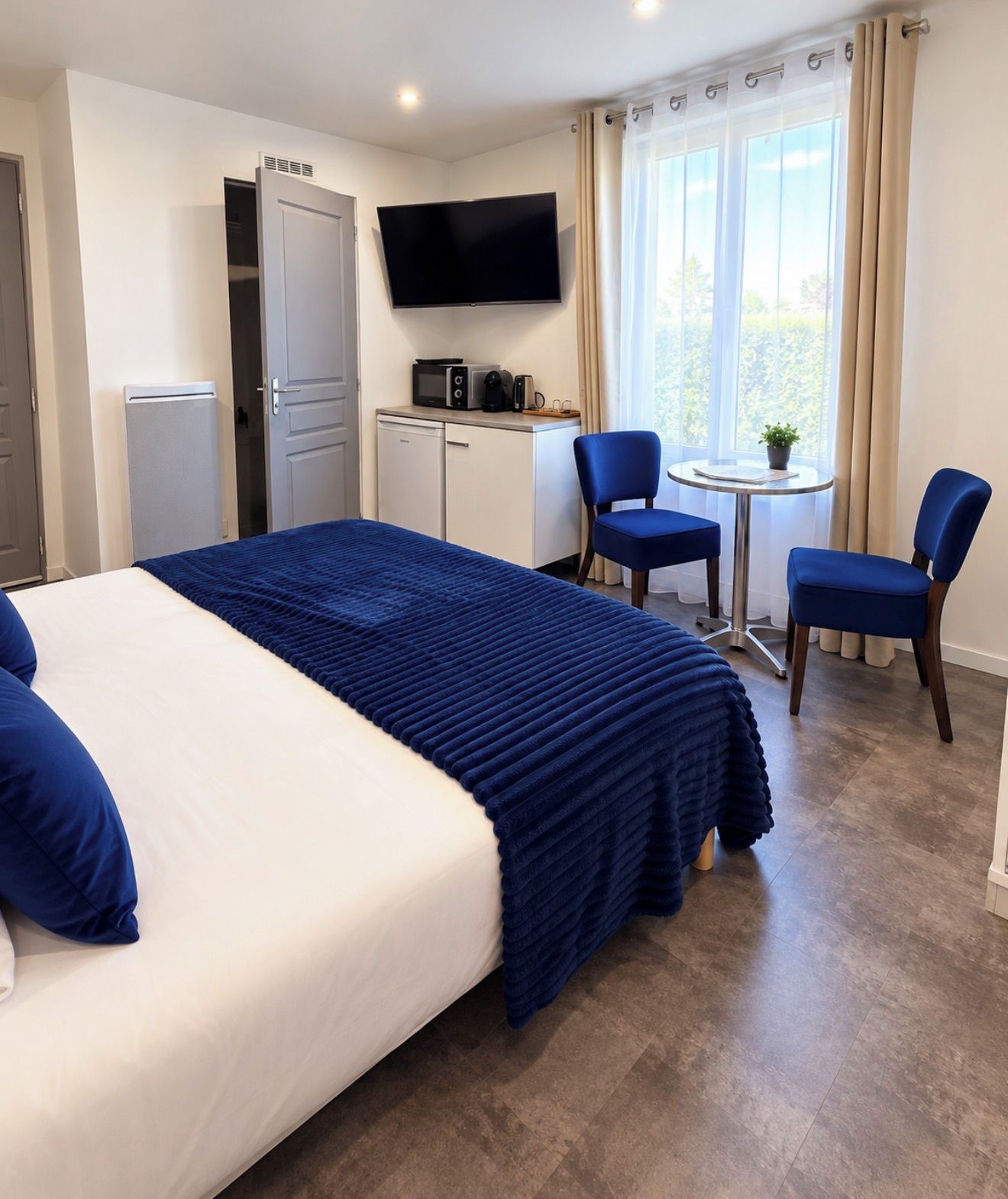 A hotel room with a large bed, a desk with a flat-screen TV, a small table with two chairs, and a window with curtains.