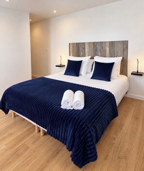 Una cama bien hecha con una cubierta azul oscuro, dos almohadas blancas y dos almohadas azul oscuro. Hay dos mesitas de noche con lámparas, y el piso es de madera.