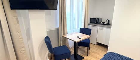 Une chambre d’hôtel avec un lit, un téléviseur fixé au mur, une petite table avec des chaises, un coin cuisine et une fenêtre avec des rideaux.