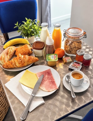 A breakfast table with croissants, cheese, ham, a bottle of Nutella, a jar of cereal, a banana, a glass of orange juice, a cup of coffee, and a plant. -  Aux Charmes (Isques)