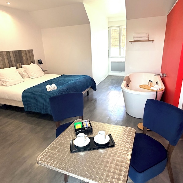 A hotel room with a bed, a sitting area with a table, a bathtub, and a window. -  Aux Charmes (Isques)