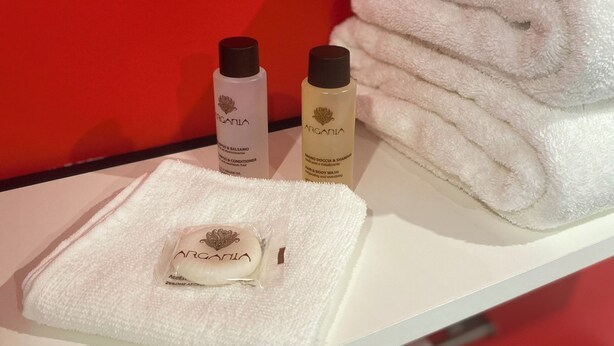 A white towel, two bottles of Argania products, and a folded towel on a white surface against a red background. -  Aux Charmes (Isques)