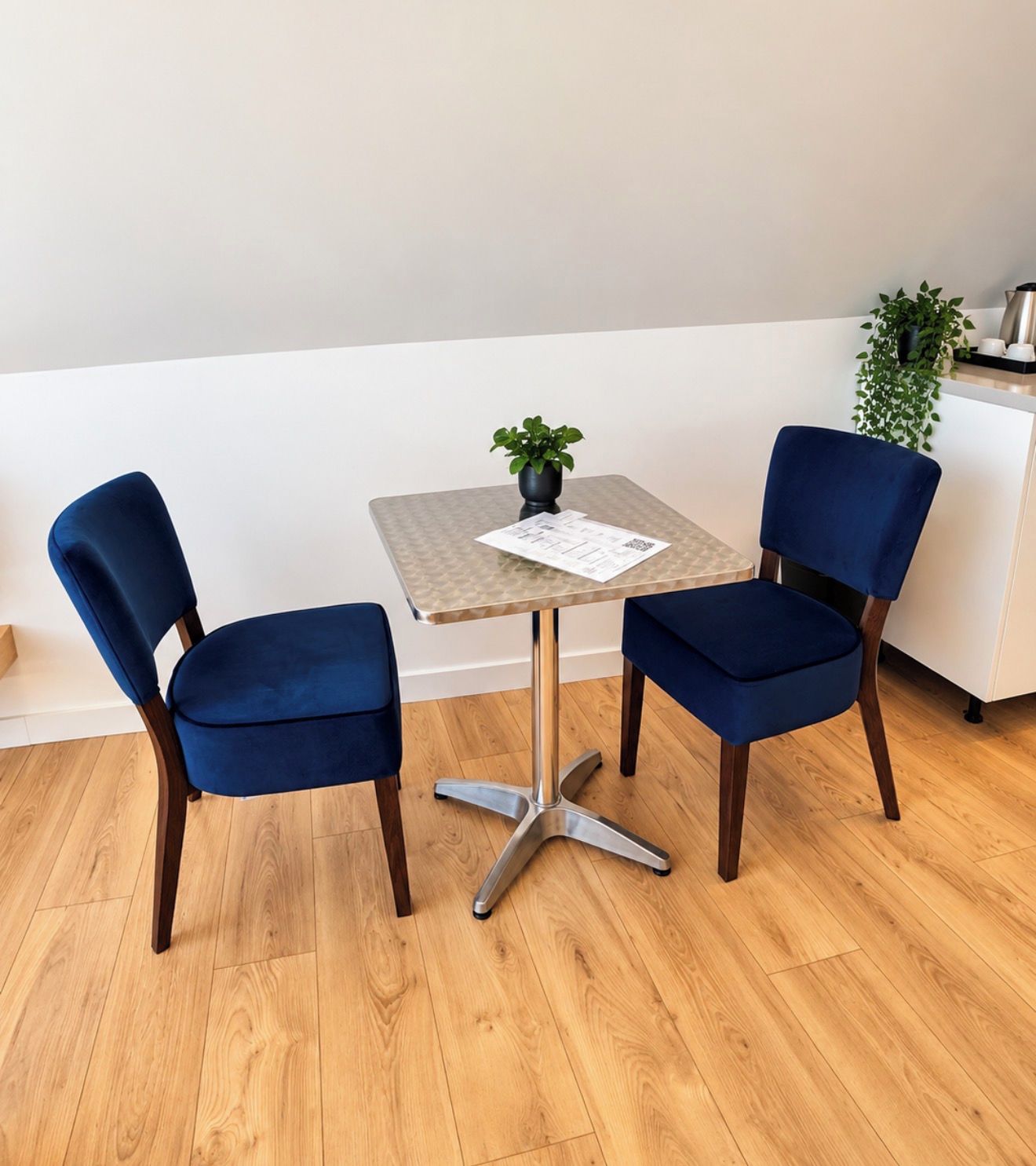 Een klein, vierkant tafeltje met een metalen onderstel en twee blauwe fauteuils. Op het tafeltje staat een potplant en er is een witte muur op de achtergrond.