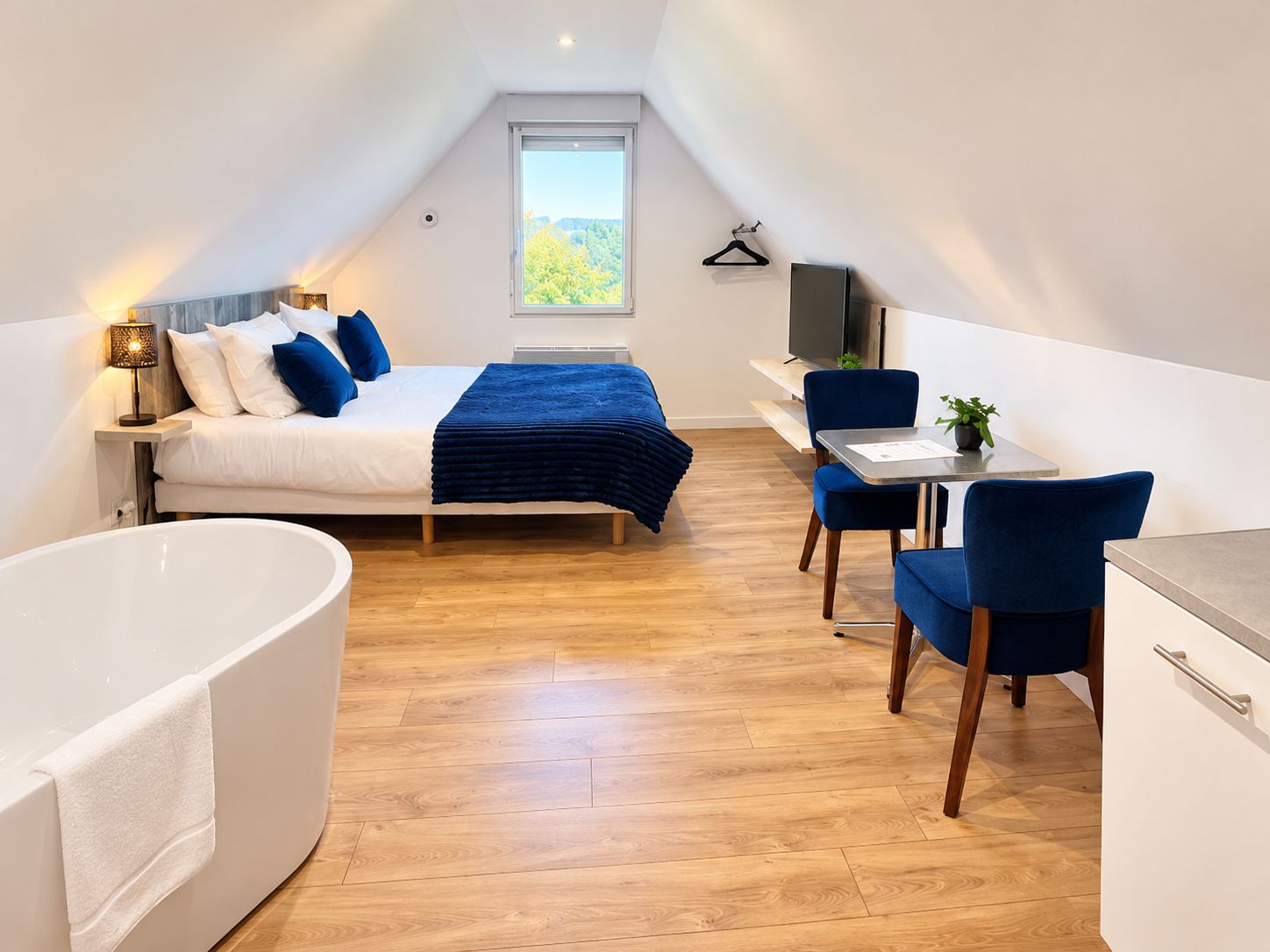 A modern bedroom with a large bed, a sitting area with a desk and chairs, and a separate bathroom with a bathtub.