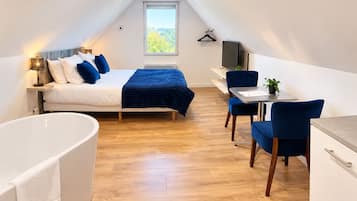 A modern bedroom with a large bed, a sitting area with a desk and chairs, and a separate bathroom with a bathtub.