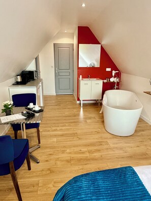 A compact room with a dining table, a blue chair, a white bathtub, and a red accent wall. -  Aux Charmes (Isques)