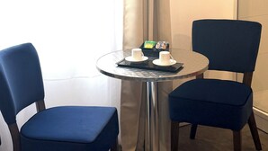 Une petite table ronde avec deux chaises, un plateau avec des tasses et une boîte de collations, et une fenêtre avec des rideaux.