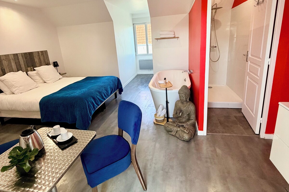 A modern hotel room with a bed, a desk with a chair, a statue, and a shower area.